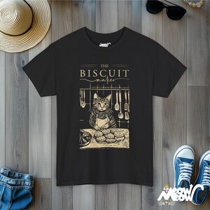 Pu&ograve; includere: Maglietta nera con una grafica di un gatto che indossa un cappello da cuoco e un grembiule, mentre fa i biscotti. Il testo "The Biscuit Maker" &egrave; sopra il gatto.