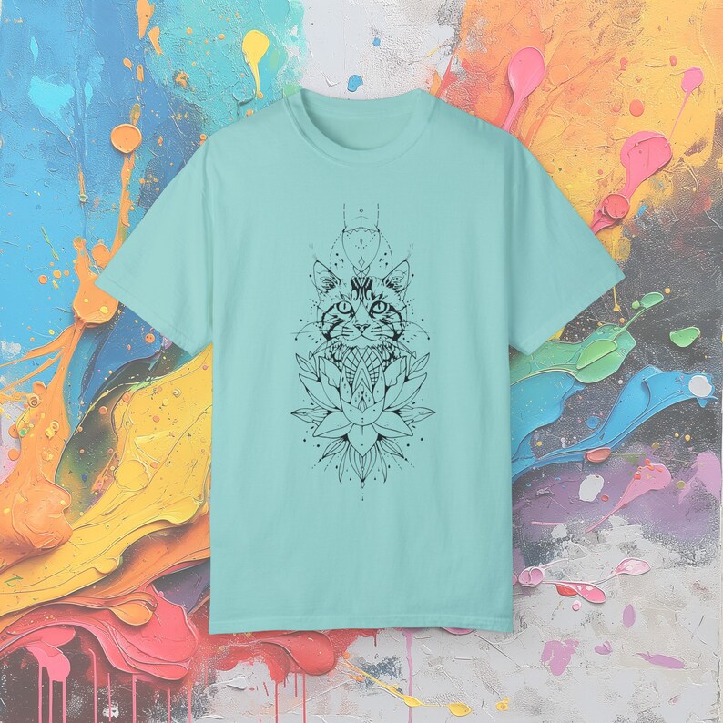 May include: A light blue t-shirt featuring a black and white graphic of a cat's face above a lotus flower. The cat design has geometric elements. The shirt is made of a soft material and has a crew neck.