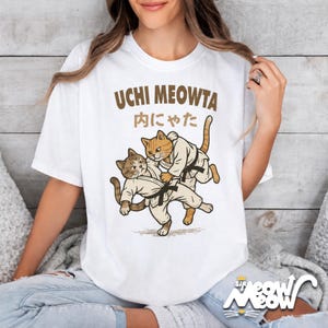 K&ouml;nnte beinhalten: Ein wei&szlig;es T-Shirt mit kurzen &Auml;rmeln und einer Grafik von zwei Katzen in wei&szlig;en Kampfkunst-Uniformen mit schwarzen G&uuml;rteln, die einen Judo-Wurf ausf&uuml;hren. &Uuml;ber den Katzen steht der Text UCHI MEOWTA in braunen Buchstaben sowie japanische Schriftzeichen.