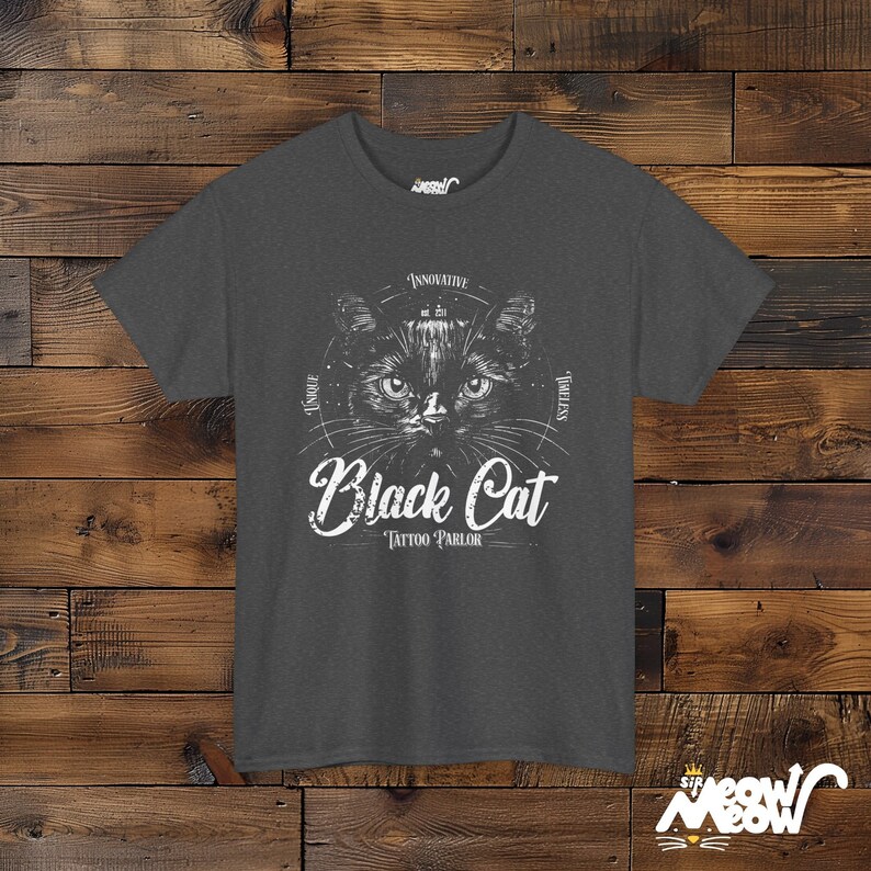 K&ouml;nnte beinhalten: Ein dunkelgraues T-Shirt mit einer schwarzen Katzenillustration und dem Text "Black Cat Tattoo Parlor" in Wei&szlig;. Die Augen der Katze sind gro&szlig; und das Design ist von einem Kreis mit den Worten "Einzigartig", "Innovativ", "Zeitlos" und "Vertrauensw&uuml;rdig" in Wei&szlig; umgeben.