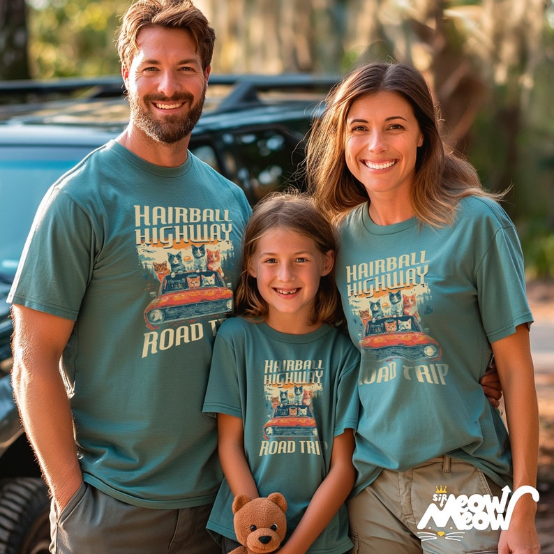 Family of a mom, dad & daughter outside. The whole family are wearing the same tshirt which features a gang of cats driving a car. Text on the shirt reads "Hairball Highway Road Trip".