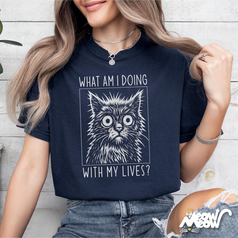 May include: Navy blue t-shirt featuring a distressed, wide-eyed cat illustration within a white rectangular frame. The text "WHAT AM I DOING WITH MY LIVES?" is above and below the cat image. The shirt is a casual wear item.