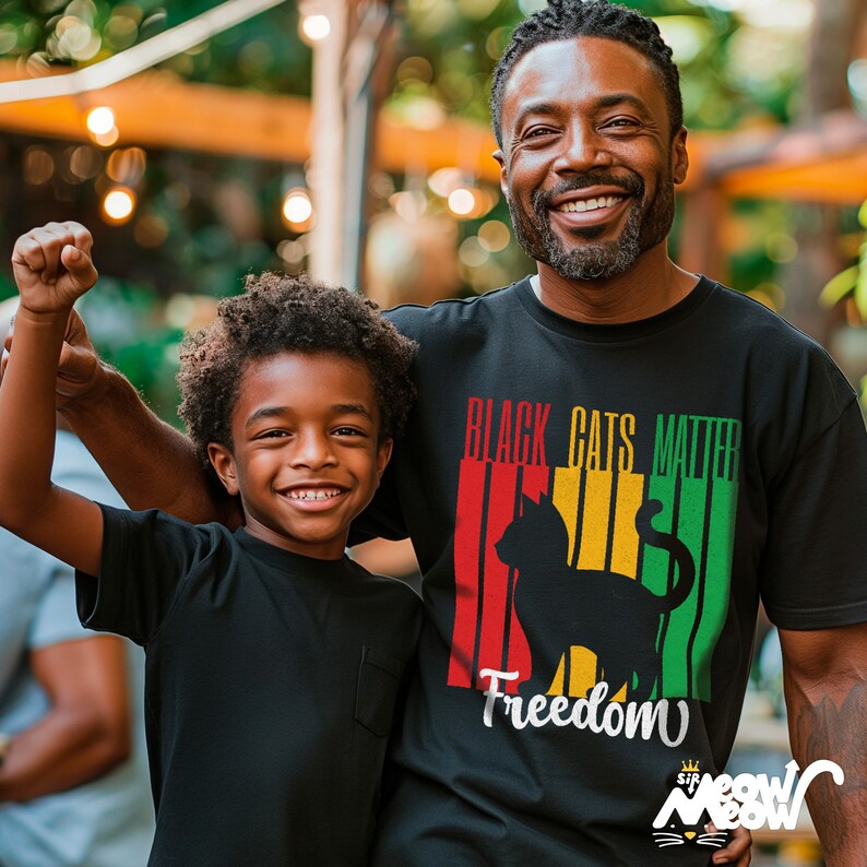 Pu&ograve; includere: Un padre e un figlio indossano magliette nere. La maglietta del padre ha una silhouette di gatto nero con il testo "Black Cats Matter" e "Freedom" in strisce rosse, gialle e verdi.