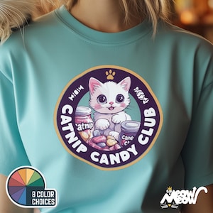 May include: A turquoise blue t-shirt with a white cat wearing a pink collar and holding a jar of catnip. The cat is surrounded by candy and the text "Meow Catnip Candy Club" in a purple circle.