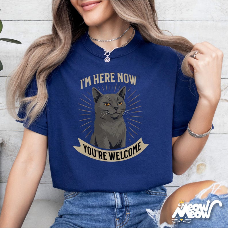 Może przedstawiać: Niebieska koszulka z ilustracją szarego kota i tekstem "I'm here now you're welcome".