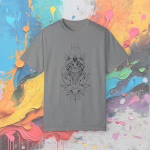 May include: A gray t-shirt featuring a black graphic of a cat's face, geometric shapes, and a lotus flower. The design is centered on the front of the shirt. The background is a colorful abstract painting.