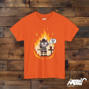 Op de afbeelding: Oranje T-shirt met een cartoonkat die een hoed draagt en op een stoel zit, omringd door vlammen. De tekst "This is fine" staat in een spraakballon boven de kat.