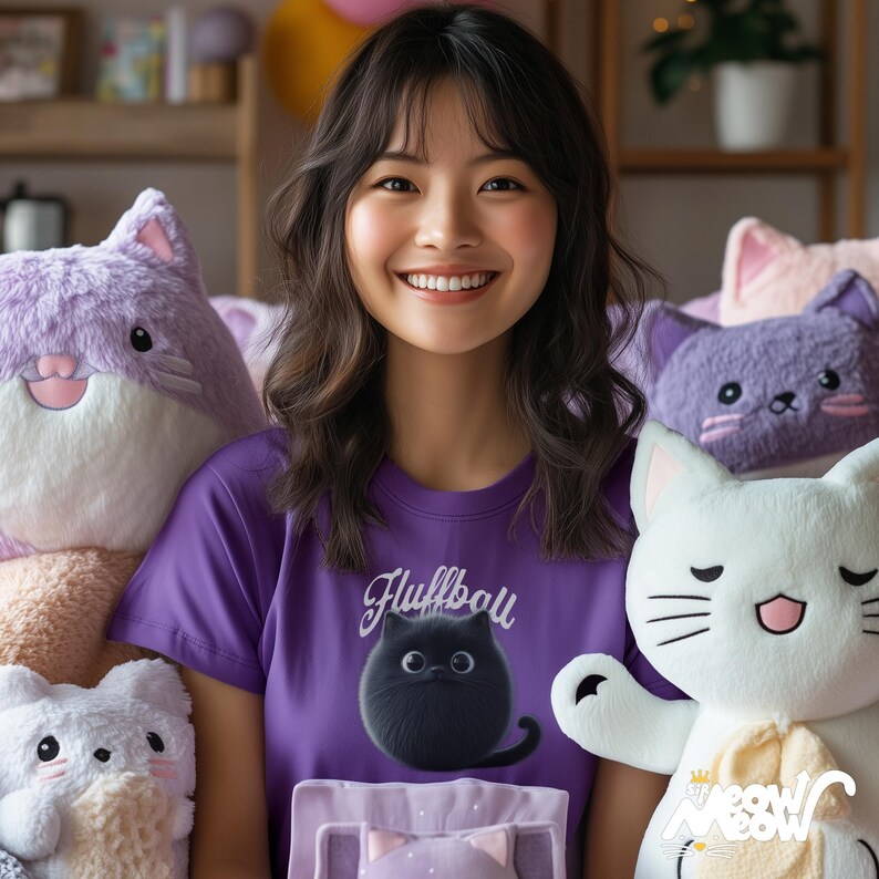 Puede incluir: Una mujer lleva una camiseta morada con el texto "Fluffball" y un gr&aacute;fico de un gato negro. Est&aacute; rodeada de peluches, incluyendo un gato morado, un gato blanco y un gato morado y blanco.