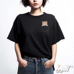 May include: Black t-shirt featuring a pocket with a cartoon cat peeking out. The cat has orange and white fur and a smiling expression. The shirt is a crew neck style and is made of a soft material.