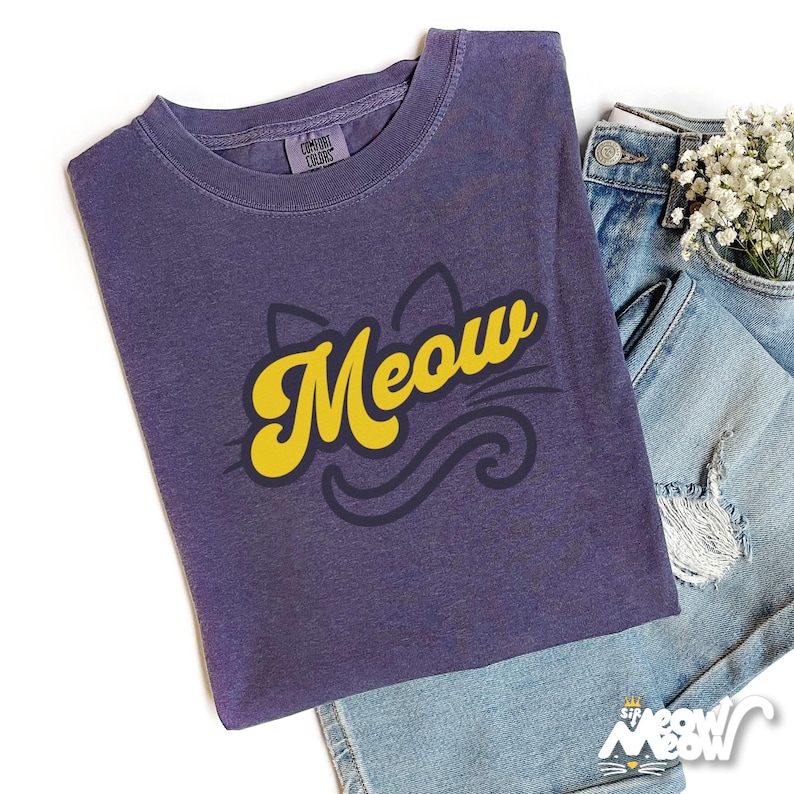 May include: A purple Comfort Colors t-shirt with a yellow "Meow" graphic and a cat silhouette. The shirt is folded on top of a pair of blue denim shorts. The shirt has a crew neck and short sleeves.