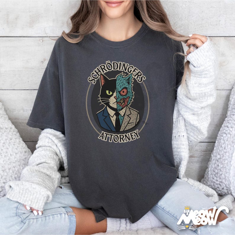 May include: A dark gray t-shirt with a graphic of a cat with a split face. One side of the face is a regular cat, and the other side is a monster. The text "Schr&ouml;dinger's Attorney" is printed below the graphic.