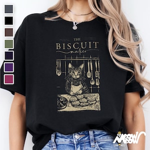 Pu&ograve; includere: Maglietta nera con una grafica di un gatto che indossa un grembiule e fa biscotti. Il testo "The Biscuit Maker" &egrave; sopra il gatto.