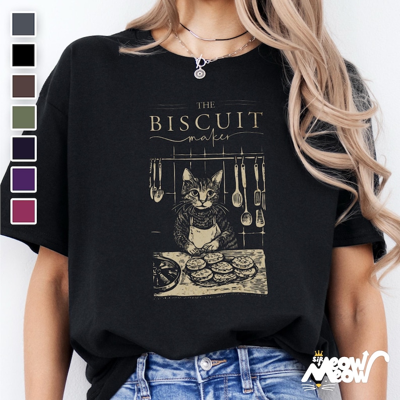 Pu&ograve; includere: Maglietta nera con una grafica di un gatto che indossa un grembiule e fa biscotti. Il testo "The Biscuit Maker" &egrave; sopra il gatto.