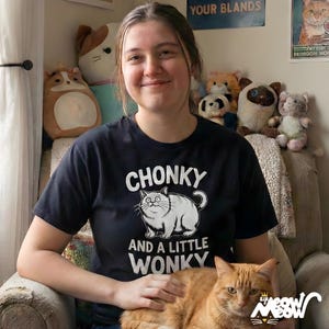 May include: A black t-shirt with the words "CHONKY AND A LITTLE WONKY" and a cartoon cat graphic. An orange cat is sitting in a person's lap. Various stuffed toys are in the background.