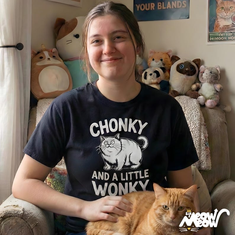 May include: A black t-shirt with the words "CHONKY AND A LITTLE WONKY" and a cartoon cat graphic. An orange cat is sitting in a person's lap. Various stuffed toys are in the background.