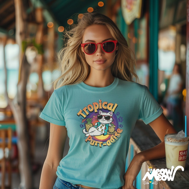 May include: A light blue t-shirt featuring a graphic of a cat wearing sunglasses and a lei, holding a pineapple drink. The graphic says "Tropical Purr-adise".