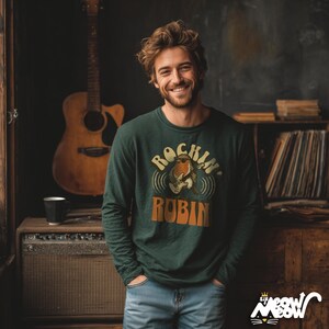 K&ouml;nnte beinhalten: Eine Person tr&auml;gt ein dunkelgr&uuml;nes Langarmshirt mit dem Text "Rockin' Robin" und einer Grafik einer Gitarre spielenden Amsel. Die Person l&auml;chelt und tr&auml;gt eine Jeans. Im Hintergrund ist eine Akustikgitarre zu sehen.