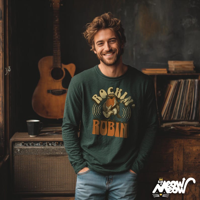 K&ouml;nnte beinhalten: Eine Person tr&auml;gt ein dunkelgr&uuml;nes Langarmshirt mit dem Text "Rockin' Robin" und einer Grafik einer Gitarre spielenden Amsel. Die Person l&auml;chelt und tr&auml;gt eine Jeans. Im Hintergrund ist eine Akustikgitarre zu sehen.