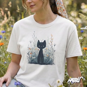 May include: Cream-colored t-shirt featuring a black cat illustration amidst wildflowers. The cat is depicted in a minimalist style, with a simple outline and large eyes. The design includes various wildflowers in shades of blue, orange, and green.