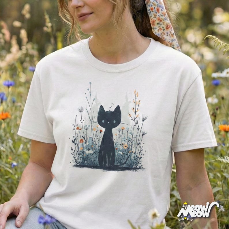 May include: Cream-colored t-shirt featuring a black cat illustration amidst wildflowers. The cat is depicted in a minimalist style, with a simple outline and large eyes. The design includes various wildflowers in shades of blue, orange, and green.