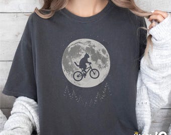 May include: Dark grey t-shirt with a cat silhouette cycling in front of a full moon. Silhouetted trees are below the moon. The shirt has the text "Sip Meow Meow" at the bottom right.