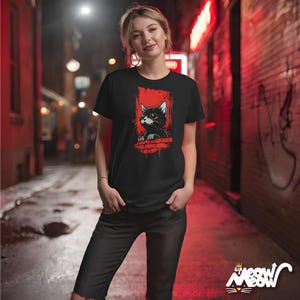 以下が含まれることがあります： レンガの壁から覗く黒猫のグラフィックが特徴の黒いTシャツ。背景は赤色です。猫は白いアクセントと大きな目をしています。シャツには、スタイリッシュなフォントで「Meow」というテキストも表示されています。