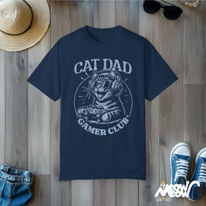 Cat Parent Gamer Club T-Shirts. The tee shows a cat holding a gaming controller. Above the image is the text "Cat Dad" and below the image is the text "Gamer Club".
The t-shirt shown is in the color "True Navy" and is also available in a "Cat Mom" version.