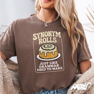 Könnte beinhalten: Braunes T-Shirt mit dem Text "SYNONYM ROLLS" über einer Abbildung einer Zimtschnecke. Darunter steht "JUST LIKE GRAMMAR USED TO MAKE". Das Shirt hat eine lockere Passform und ist aus weichem Material.