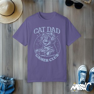 Cat Parent Gamer Club T-Shirts. The tee shows a cat holding a gaming controller. Above the image is the text "Cat Dad" and below the image is the text "Gamer Club".
The t-shirt shown is in the color "Grape" and is also available in a "Cat Mom" version.