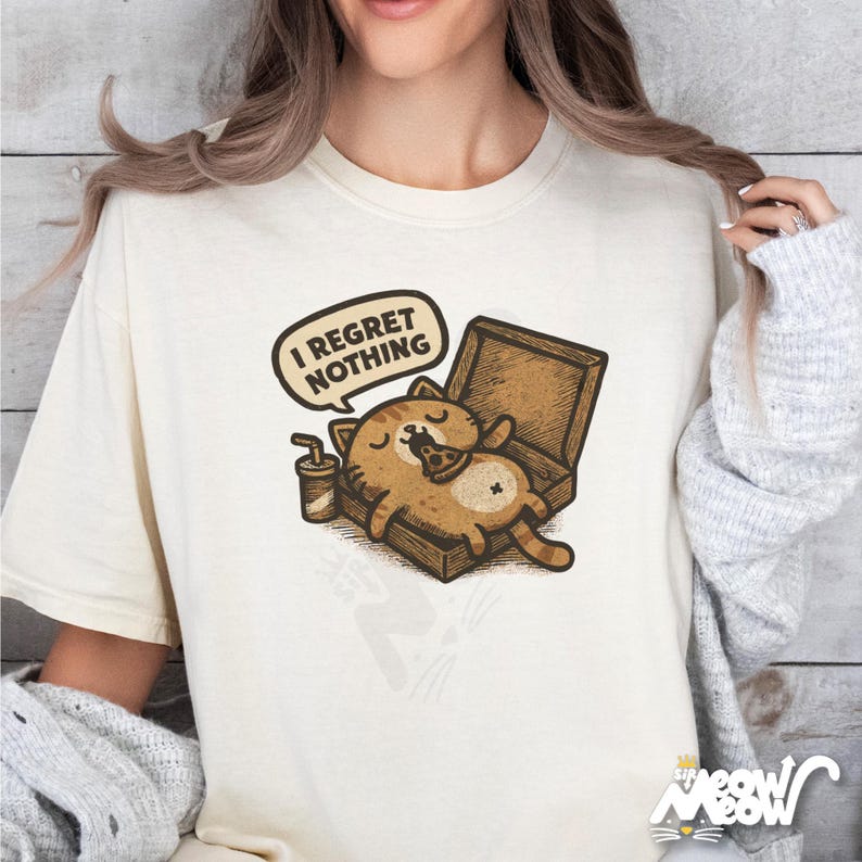 K&ouml;nnte beinhalten: Cremefarbenes T-Shirt mit einer Cartoon-Katze in einer Pizzaschachtel, die Pizza isst. Eine Sprechblase &uuml;ber der Katze lautet "I REGRET NOTHING". Ein kleines Getr&auml;nk mit Strohhalm befindet sich neben der Katze. Das Shirt hat eine lockere Passform.