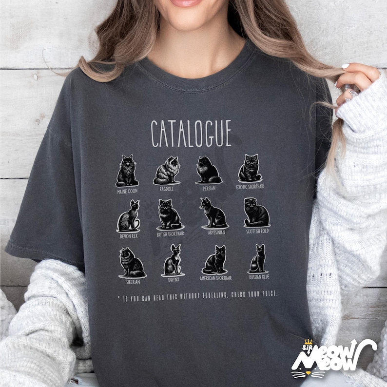 May include: Dark gray t-shirt featuring a "CATALOGUE" design with illustrations of various cat breeds, including Maine Coon, Ragdoll, Persian, and Exotic Shorthair. The shirt also includes the text: "IF YOU CAN READ THIS WITHOUT SQUINTING, CHECK YOUR PRIZE."
