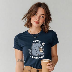 May include: Navy blue t-shirt featuring a cartoon cat holding a coffee cup, with the text "SSHH... COFFEE FIRST". The cat is gray with a white steam cloud. The shirt is a crew neck and is made of a soft material.