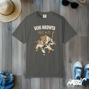 K&ouml;nnte beinhalten: Ein anthrazitfarbenes T-Shirt mit einer Grafik von zwei Katzen in wei&szlig;en Kampfsportanz&uuml;gen mit schwarzen G&uuml;rteln, die einen Judowurf ausf&uuml;hren. Der Text UCHI MEOWTA und japanische Schriftzeichen sind in Wei&szlig; und Beige dar&uuml;ber gedruckt.