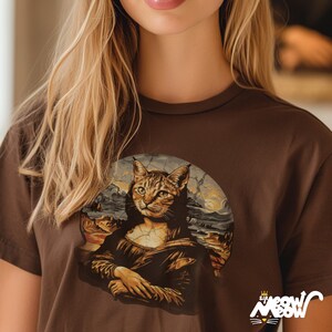Peut inclure: T-shirt marron avec un portrait de chat inspir&eacute; de la Joconde. Le visage du chat est au centre, avec un paysage en arri&egrave;re-plan. Le t-shirt porte l'inscription "Meow Meow" dans une police stylis&eacute;e.
