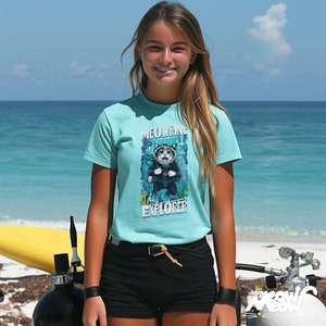 May include: A young person wearing a turquoise blue t-shirt with a graphic of a cat wearing scuba gear and the text "MEOWRING EXPLORER". The person is standing on a beach with a surfboard and scuba gear in the background.
