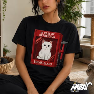 May include: Black t-shirt featuring a red graphic of a fire alarm box with a white cat inside. The text on the box reads "IN CASE OF DEPRESSION BREAK GLASS". A hammer is attached to the side of the box.