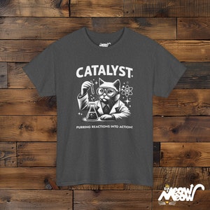 May include: A dark gray t-shirt with a white graphic of a cat wearing glasses and a lab coat holding a test tube and beaker. The text "CATALYST" is above the cat and "PURRING REACTIONS INTO ACTION!" is below the cat.