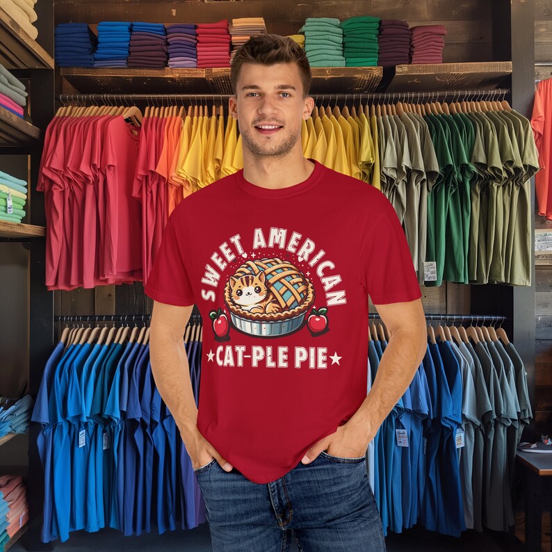 May include: Red t-shirt with a white graphic of a cat inside a pie. The text on the shirt reads "Sweet American *Cat-ple Pie*"