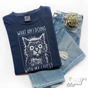 May include: Navy blue t-shirt with a distressed cat graphic and the text "WHAT AM I DOING WITH MY LIVES?". The shirt is paired with light blue denim shorts and a sprig of white flowers.