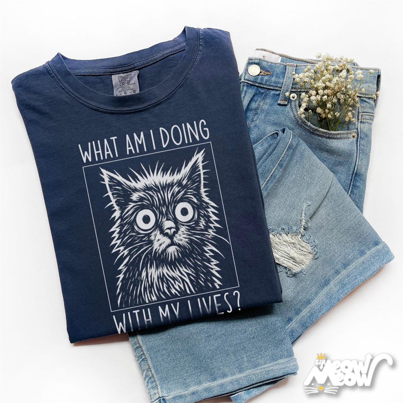 May include: Navy blue t-shirt with a distressed cat graphic and the text "WHAT AM I DOING WITH MY LIVES?". The shirt is paired with light blue denim shorts and a sprig of white flowers.