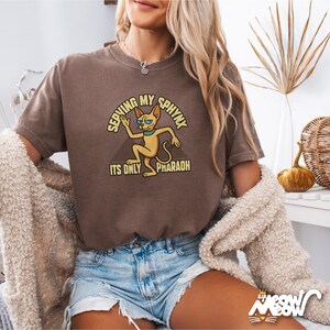 May include: A brown t-shirt featuring a cartoon sphinx cat wearing sunglasses and the text "Serving My Sphinx It's Only Pharaoh".