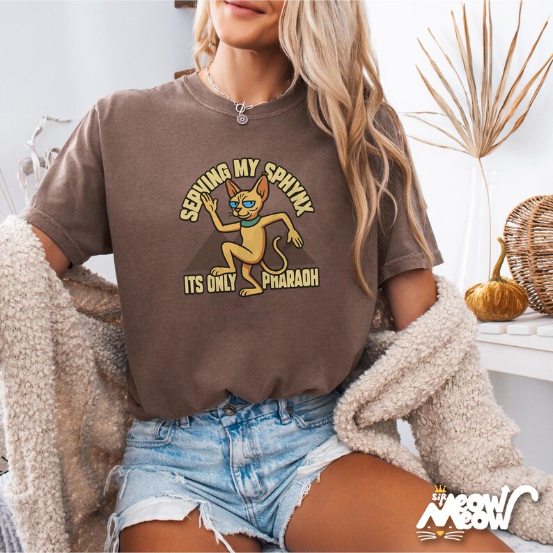 May include: A brown t-shirt featuring a cartoon sphinx cat wearing sunglasses and the text "Serving My Sphinx It's Only Pharaoh".