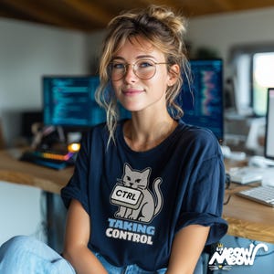 K&ouml;nnte beinhalten: Marineblaues T-Shirt mit einer Grafik einer grauen Katze, die eine "CTRL"-Taste h&auml;lt, darunter der Text "TAKING CONTROL". Das Shirt geh&ouml;rt zur "Sir Meow Meow" Kollektion. Die Person im Bild tr&auml;gt eine Brille und hat blondes Haar.