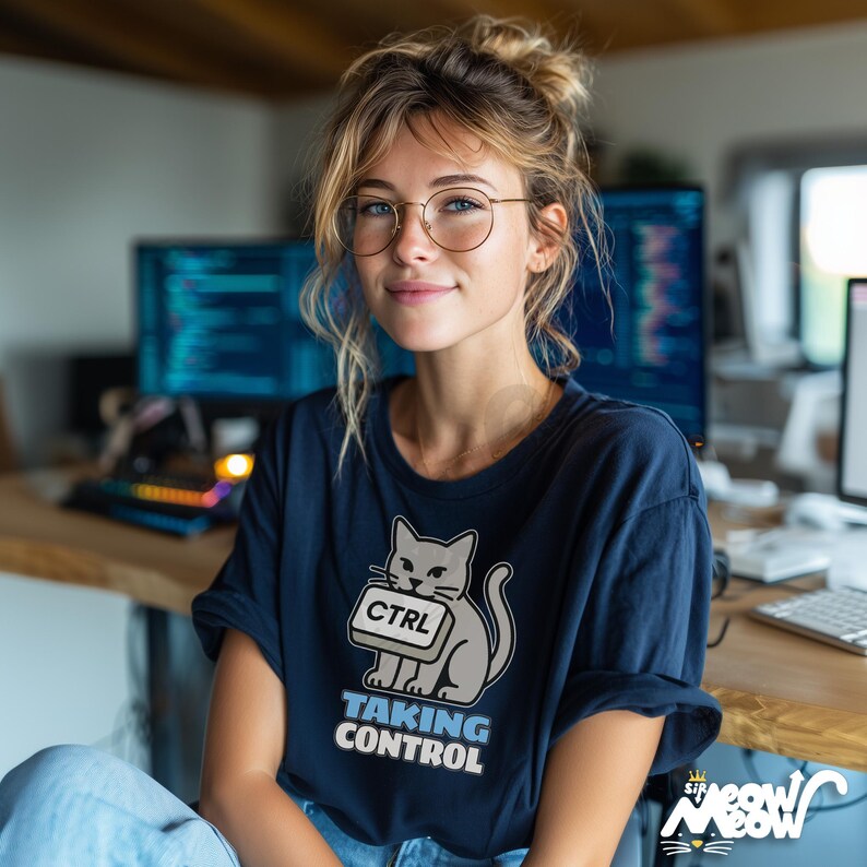 K&ouml;nnte beinhalten: Marineblaues T-Shirt mit einer Grafik einer grauen Katze, die eine "CTRL"-Taste h&auml;lt, darunter der Text "TAKING CONTROL". Das Shirt geh&ouml;rt zur "Sir Meow Meow" Kollektion. Die Person im Bild tr&auml;gt eine Brille und hat blondes Haar.