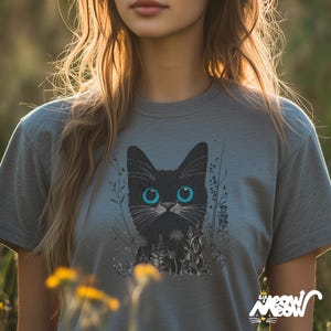May include: A gray t-shirt with a black cat with blue eyes printed on the front. The cat is surrounded by black and white foliage.