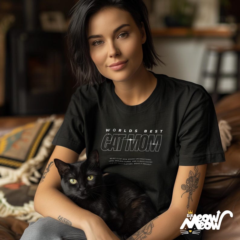 May include: A woman wearing a black t-shirt with the text "Worlds Best Cat Mom" in white. She is holding a black cat in her lap. The text on the shirt reads "Being a cat mom means unconditional love, endless purrs, and always having someone to cuddle. You love cat clothes, wear it proudly."