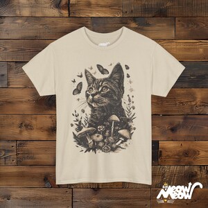Vintage style t-shirt print of a cat in the woodlands surrounded by wildflowers, mushrooms and butterflies. Sand colored tee.