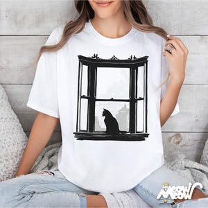 May include: White t-shirt featuring a black graphic of a cat silhouette sitting in a bay window. The window design includes architectural details and a cityscape view. The shirt has a crew neck and short sleeves.