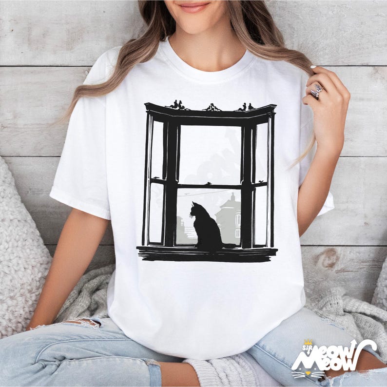 May include: White t-shirt featuring a black graphic of a cat silhouette sitting in a bay window. The window design includes architectural details and a cityscape view. The shirt has a crew neck and short sleeves.