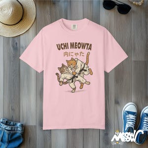 K&ouml;nnte beinhalten: Ein hellrosa T-Shirt mit kurzen &Auml;rmeln und einer Grafik von zwei Katzen in wei&szlig;en Kampfkunstanz&uuml;gen mit schwarzen G&uuml;rteln, die einen Judowurf ausf&uuml;hren. &Uuml;ber den Katzen steht der Text UCHI MEOWTA und japanische Schriftzeichen.